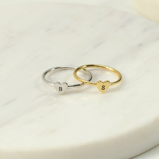 Two rings, one with a heart-shaped initial in gold colour and another in silver colour, placed on a marble surface.