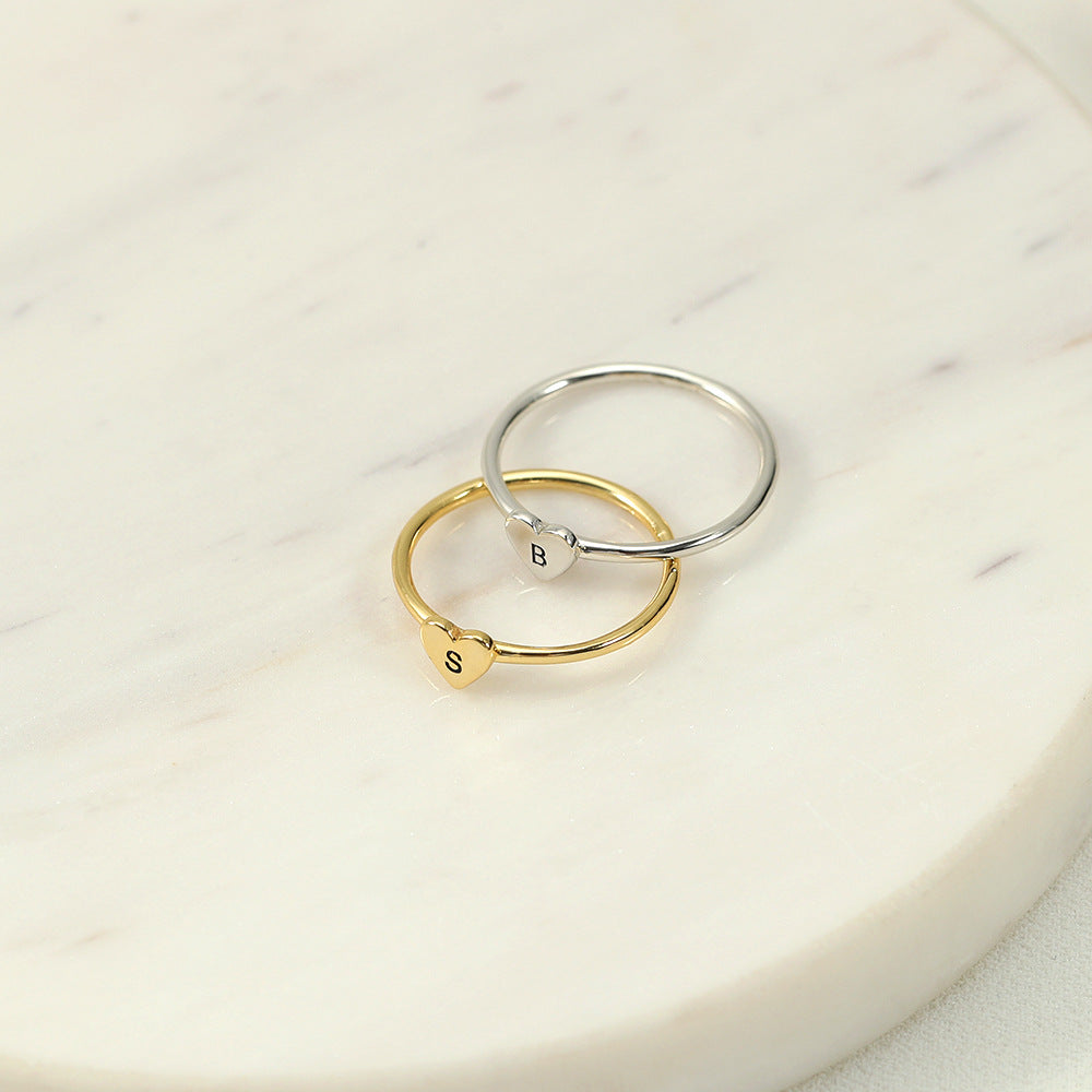 Two rings, one with a heart-shaped initial in gold colour and another in silver colour, placed on a marble surface.