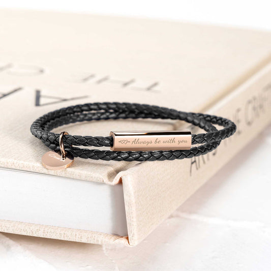 A black leather wrap bracelet with a stainless steel tag lying on an open book, with the tag displaying an engraving.