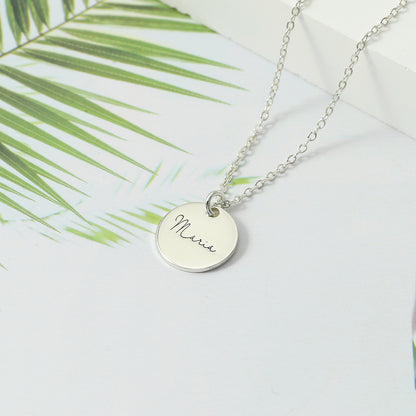 Silver disc necklace pendant displayed on a white background with palm leaves in the background.