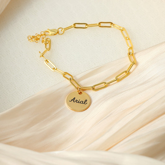 A gold-coloured bracelet with a round disc charm personalised with the name 'Arial'.