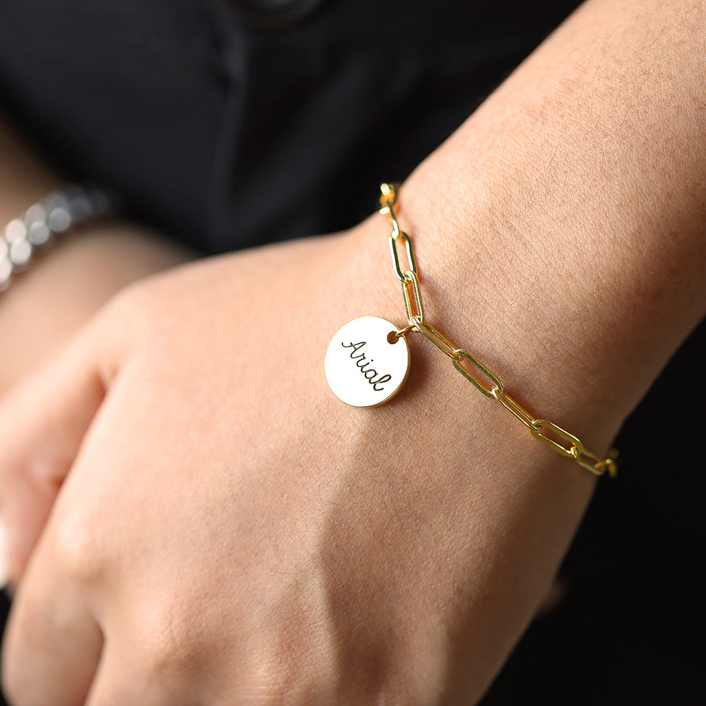 A gold-coloured bracelet with a round disc charm personalised with the name 'Arial'.