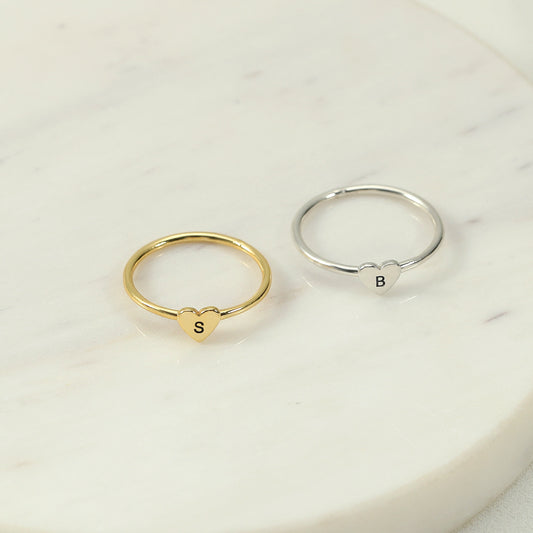 Two rings, one with a heart-shaped initial in gold colour and another in silver colour, placed on a marble surface.
