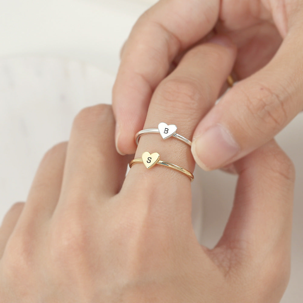 Two rings, one with a heart-shaped initial in gold color and another in silver color, on an index finger.