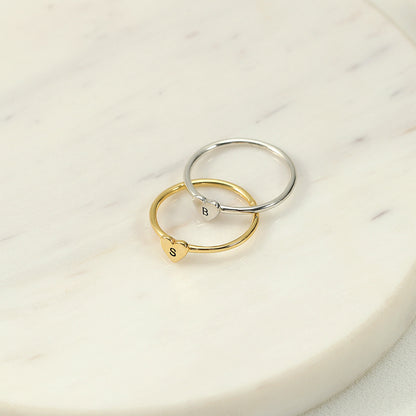 Two rings, one with a heart-shaped initial in gold colour and another in silver colour, placed on a marble surface.