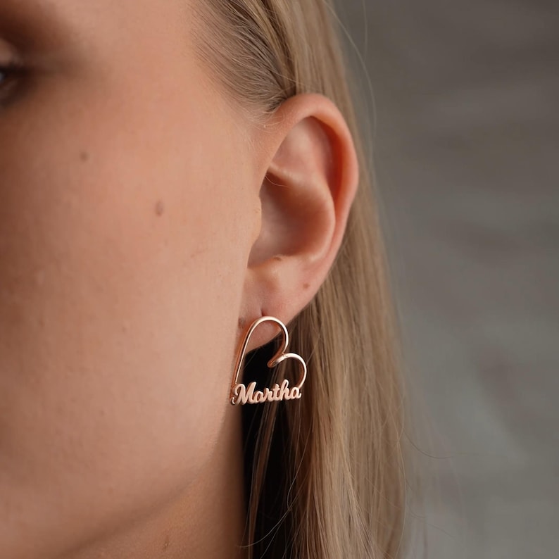 Close-up of a person wearing gold heart-shaped earrings with 'Martha' engraving.