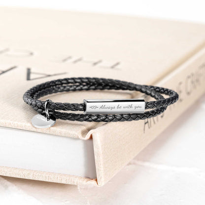A black leather wrap bracelet with a stainless steel tag lying on an open book, with the tag displaying an engraving.