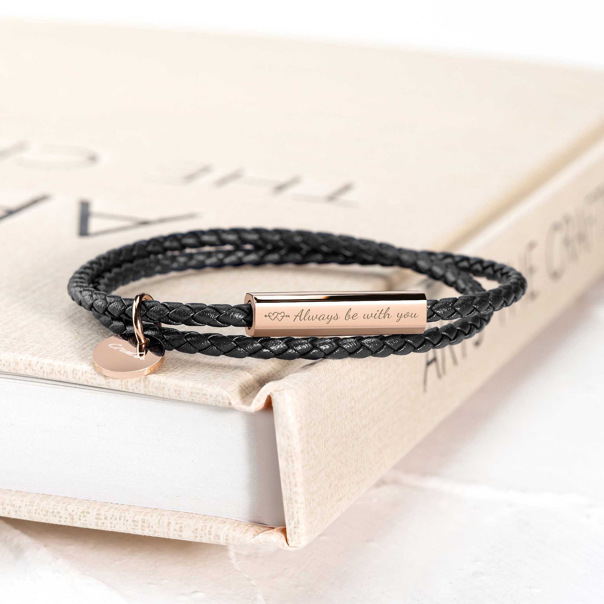 A black leather wrap bracelet with a stainless steel tag lying on an open book, with the tag displaying an engraving.