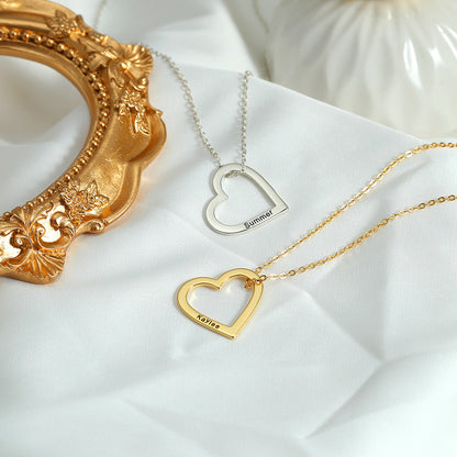 Two necklaces, one with a gold heart and the other with a silver heart, each customised with different names, lying on a white surface with a golden ornament in the background.