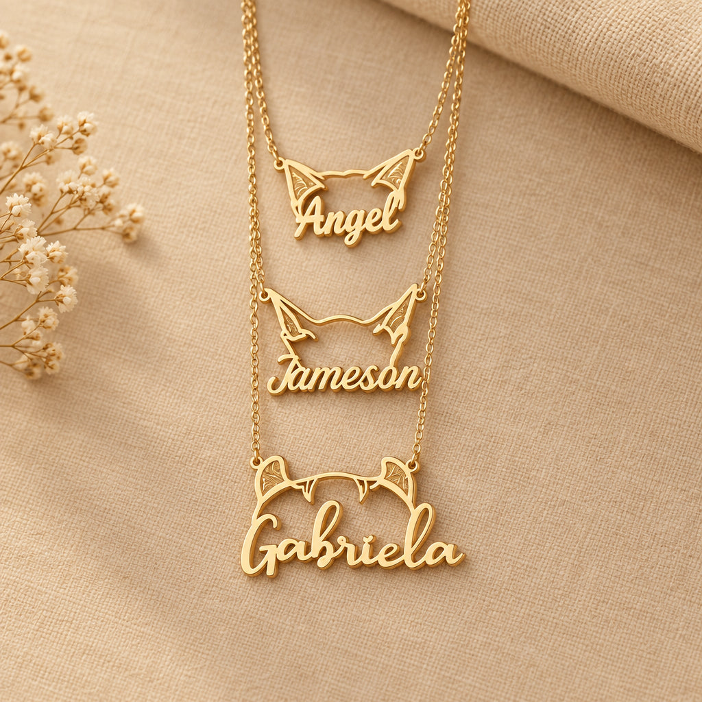 Gold name necklaces on beige material with cat ears and 'Angel', 'Jameson', and 'Gabriela' inscribed.