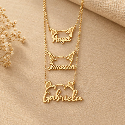 Gold name necklaces on beige material with cat ears and 'Angel', 'Jameson', and 'Gabriela' inscribed.