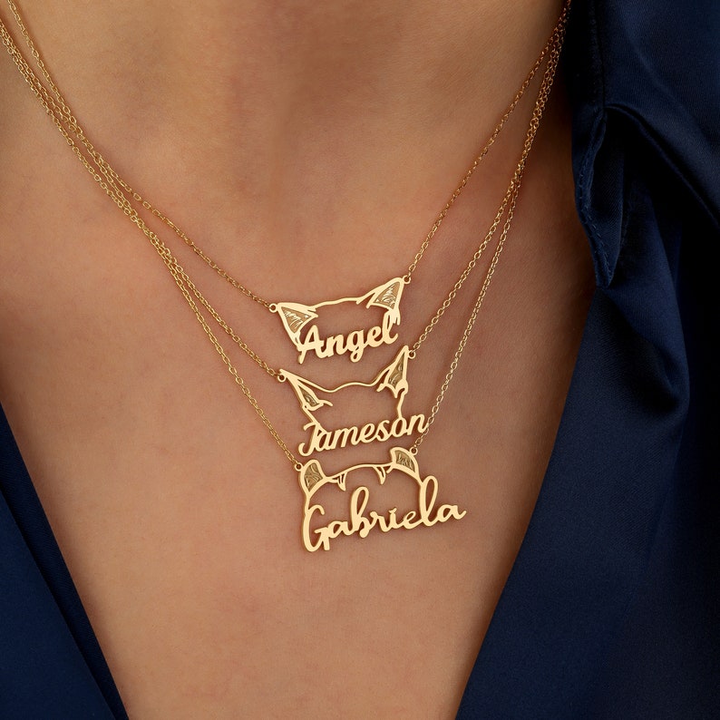Gold name cat ear necklace on a person's neck with 'Angel', 'Jameson', and 'Gabriela' inscribed.