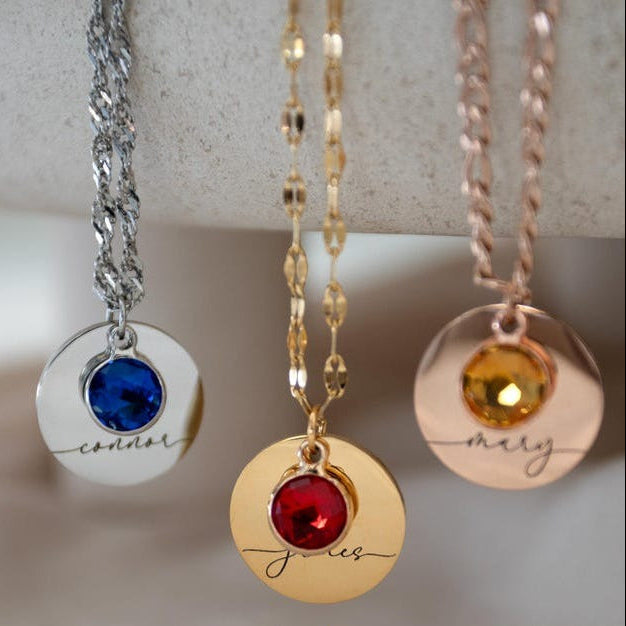Three necklaces with round pendants featuring names and colored stones on a neutral background.