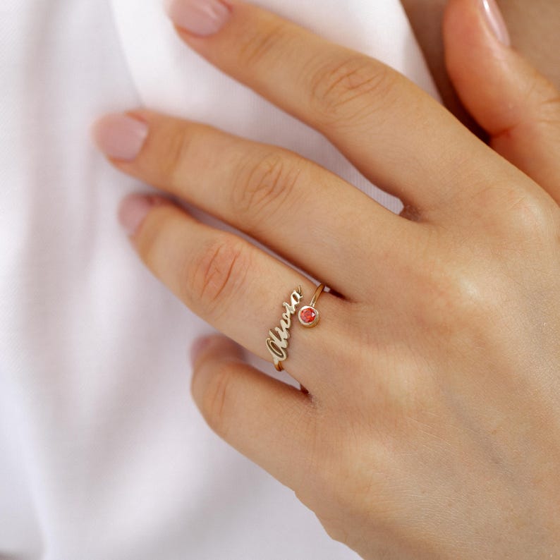 Hand wearing a gold ring with 'Olivia' and a red gemstone on a white background
