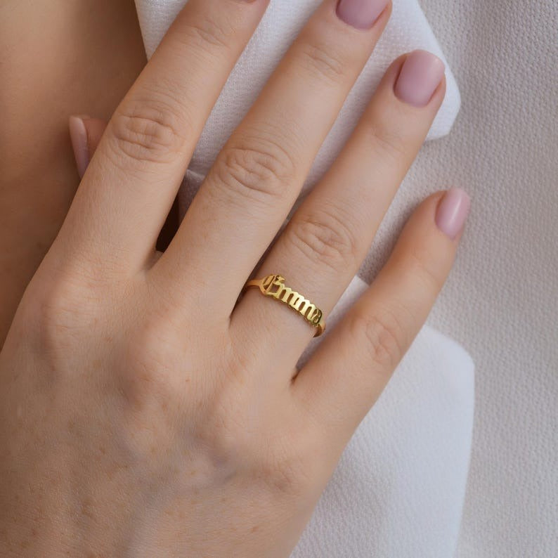 Hand wearing a gold name ring on a neutral background
