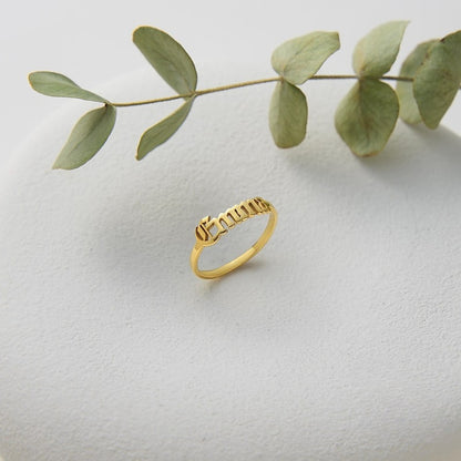 Gold name ring on a light gray surface with a branch of eucalyptus leaves.